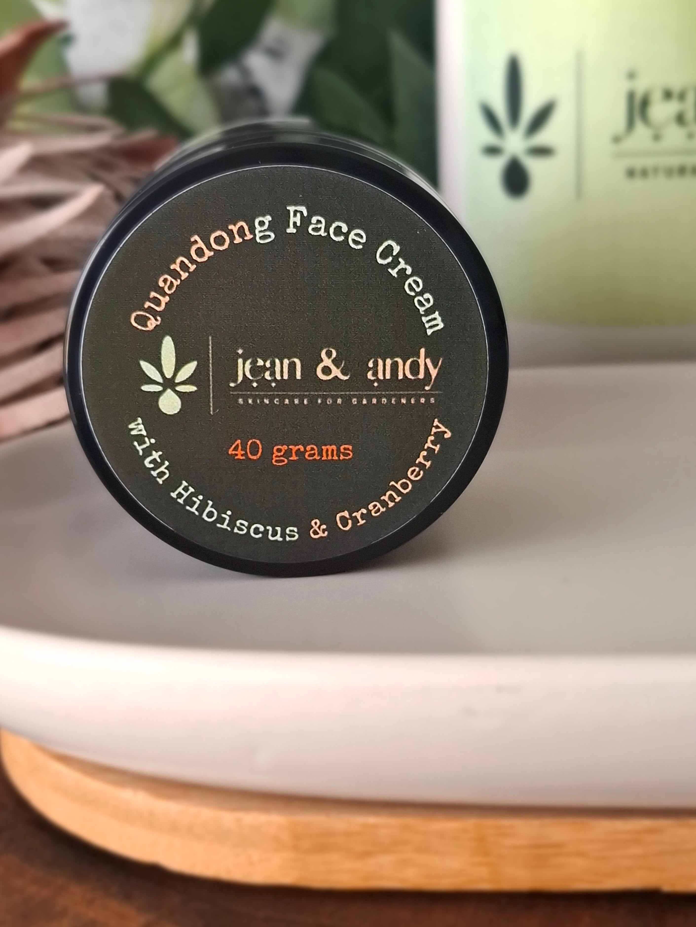 Close-up of a round container of Quandong Face Cream with Hibiscus & Cranberry by jean & andy on a white dish with a wooden base. The label indicates 40 grams and highlights ingredients such as cranberry seed oil and hibiscus. A blurred plant in the background adds a natural touch.