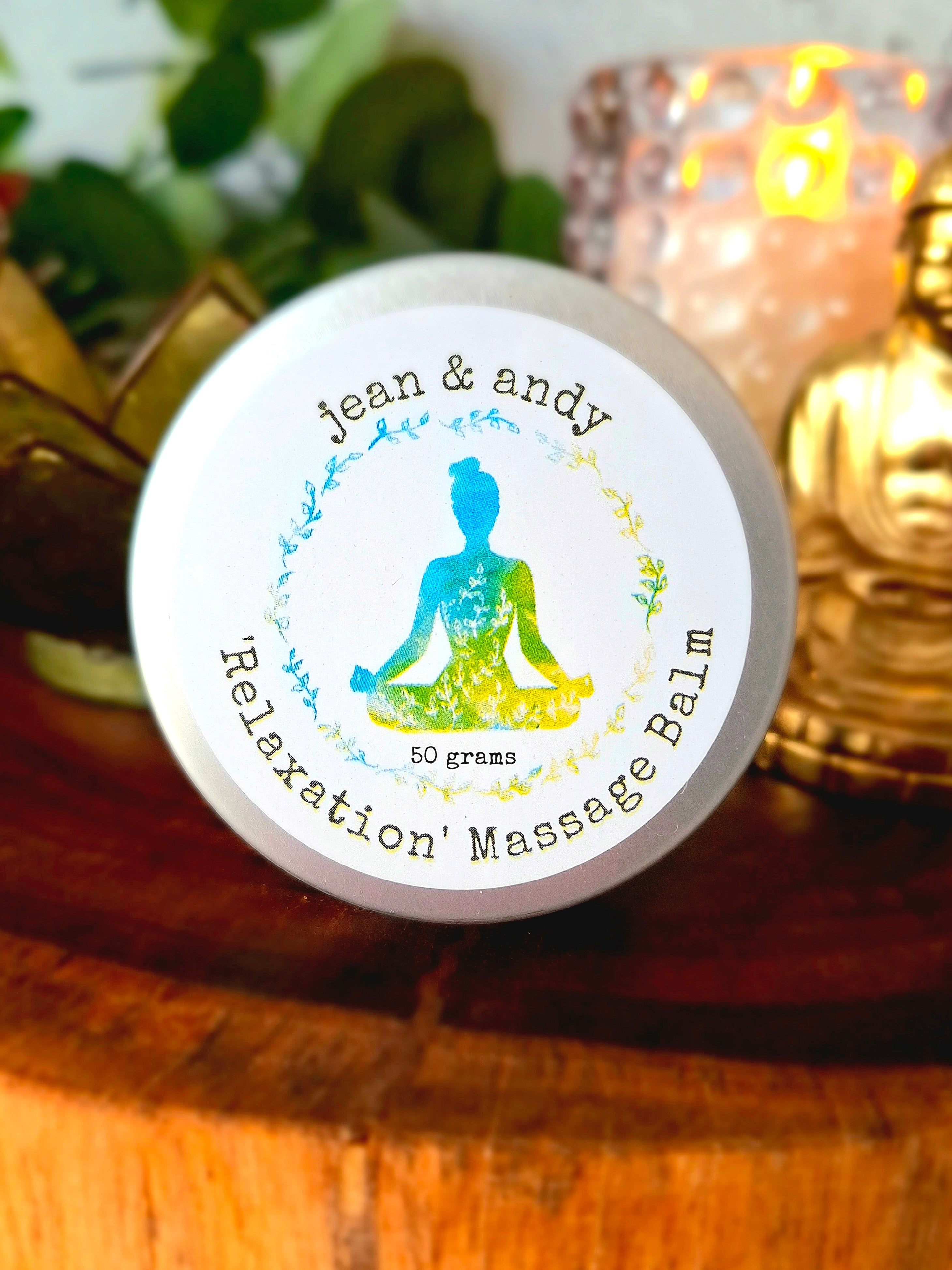 A tin of Relaxation Massage Balm by jean & andy is placed on a wooden surface. The label features a colorful silhouette of a meditating person surrounded by a leafy wreath. Infused with calming essential oils, the tin enhances the serene ambiance alongside candles and plants.