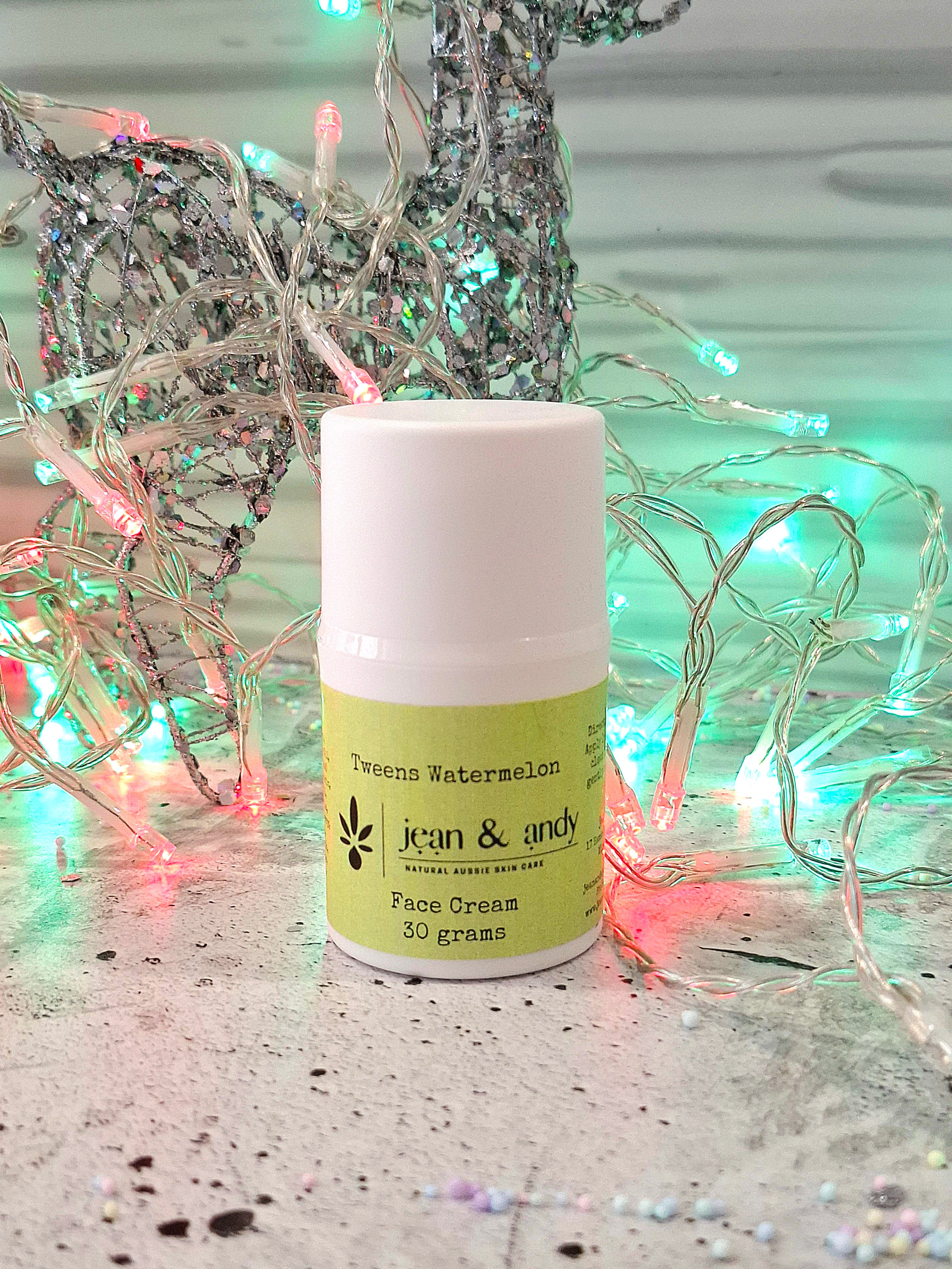 A small jar of jean & andy Tweens Face Lotion with Watermelon & Raspberry Seed rests on a surface surrounded by colorful fairy lights. The jar is white with a green label, containing 30 grams of product. In the background, a decorative silver reindeer adds a festive touch.