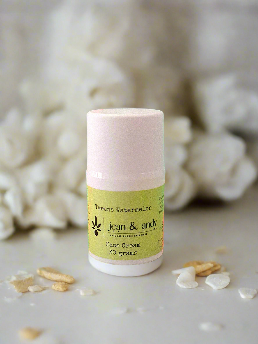 A 30-gram container of Tweens Face Lotion with Watermelon & Raspberry Seed by jean & andy is nestled among white stones and scattered petals. The lotion comes in a cylindrical container with a light pink lid and a green label.